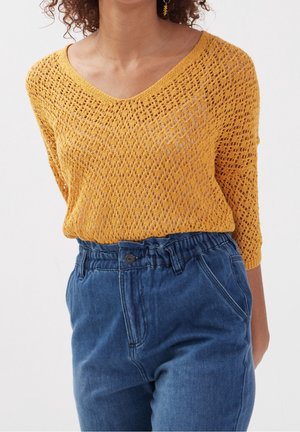 Femme portant un pull en maille ajourée jaune moutarde à manches 3/4, associé à un pantalon en denim bleu taille haute avec une ceinture froncée.