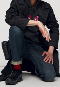 Abrigo negro largo sobre una sudadera gris con logo rojo, jeans azules con los bajos enrollados, zapatillas negras y un pequeño bolso negro.