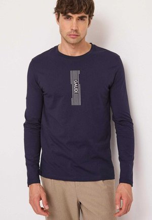 Giovane uomo che indossa una camicia blu navy a maniche lunghe con strisce bianche verticali e testo "GAUDI", abbinata a pantaloni beige, in piedi davanti a uno sfondo bianco.