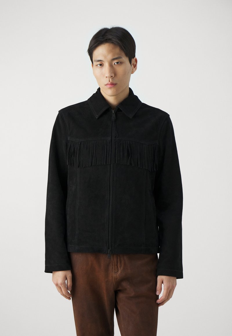 Veste en suede noire avec une fermeture éclair à l'avant, dotée d'un détail de franges sur la poitrine. Associée à un pantalon texturé marron.