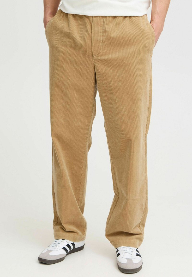 Beige Cordhosen mit entspannter Passform, ausgestattet mit einem elastischen Bund und Seitentaschen; kombiniert mit weißen und schwarzen Sneakers.