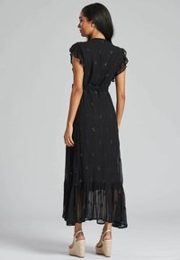 Robe maxi noire à manches courtes froncées, avec une texture transparente et des broderies en forme de diamant. Portée avec des sandales à talons compensés beiges.