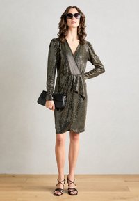 Robe enveloppante pailletée en doré métallique et noir avec des manches longues, une taille froncée et un décolleté en V profond. Pochette noire et escarpins à lanières.