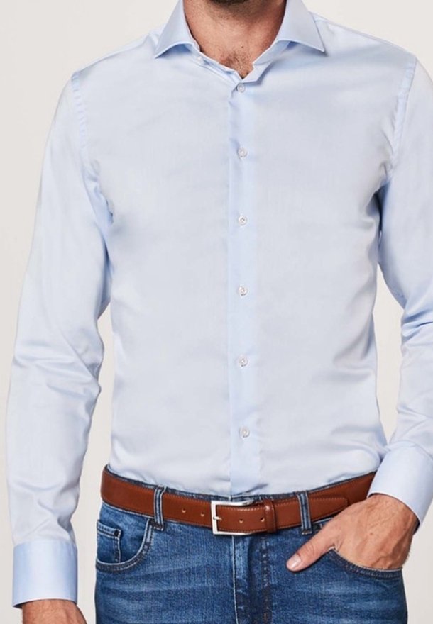 Homme portant une chemise bleu clair boutonnée rentrée dans un jean bleu avec une ceinture en cuir marron, main reposant dans sa poche.