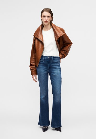 Καφέ δερμάτινο μπουφάν με φαρδύ γιακά, λευκή μπλούζα και σκούρα μπλε τζιν με φαρδιά άκρα. Το μοντέλο ποζάρει με τα χέρια στη μέση, φορώντας μαύρα μυτερά παπούτσια.