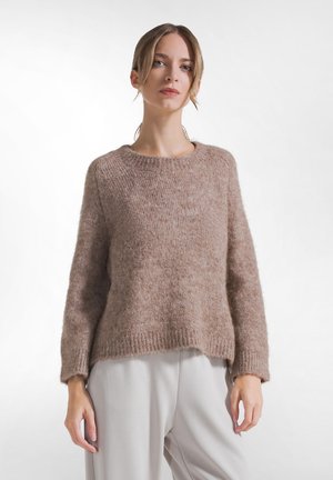 Kvinde iført en blød brun strikket sweater og lysegrå bukser, stående mod en ensfarvet hvid baggrund.