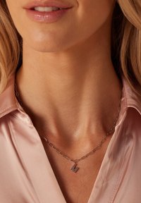 Zilveren ketting met paperclip schakels en een kleine "M" hanger, gedragen over een lichtroze satijnen blouse. Gladde textuur en minimalistisch ontwerp.