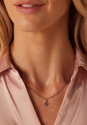 Collier en argent avec des maillons en forme de trombone et un petit pendentif "M", porté sur un chemisier en satin rose clair. Texture lisse et design minimaliste.
