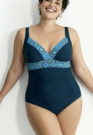 Femme souriante portant un maillot de bain une pièce bleu foncé avec des bordures bleues à motifs au niveau de la poitrine et de la taille, debout sur un fond blanc.