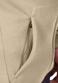 Tessuto di felpa beige con una superficie liscia, dotato di una tasca laterale con zip per il riposo. La zip ha un cursore metallico scuro.