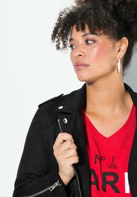 Black suede moto jacket with silver hardware, layered over a red V-neck shirt featuring a graphic design. Curly hair and hoop earring accent.