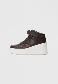 Donkerbruin hoge sneaker met dikke witte wigzool, klittenbandsluiting en veters, weergegeven in zijprofiel op een witte achtergrond.