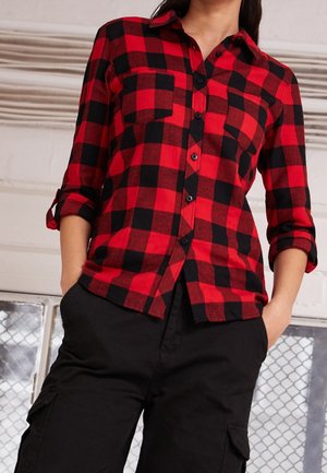 Person wearing a red and black checkered button-up shirt with rolled sleeves and black cargo pants, standing with hands in pockets.