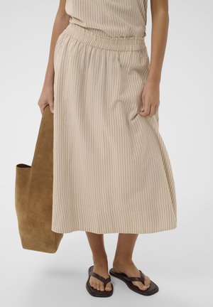 Kobieta ubrana w beżową spódnicę midi w paski i czarne klapki, trzyma dużą brązową zamszową torbę typu tote przy boku.