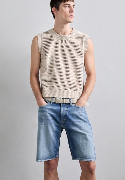 Top fără mâneci, tricotat, de culoare bej, cu un model texturat, asociat cu pantaloni scurți din denim de culoare bleu deschis. Ținuta include o curea asortată.