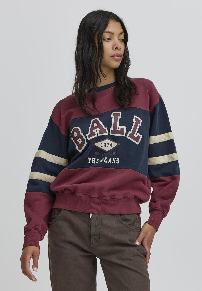 Bordo ja tumesinine ülev suurusest sviiter, millel on "BALL" graafika, soonikkraed ja horisontaalsed triibud varrukatel. Kombineeritud pruuni pükstega.