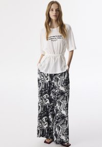 Witte oversized shirt met zwarte tekst, cinched in de taille met een strik. Zwart-wit gestreepte wijde broek met intrigerende patronen.