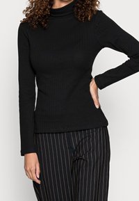 Mustassa ribattussa poolopaidassa on pitkät hihat, tiukka malli, valmistettu joustavasta kankaasta, ja sitä on yhdistetty raidallisiin mustiin housuihin.