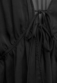 Gros plan sur une robe en tissu noir montrant une taille froncée et un détail de nœud noué sur une fine bretelle.