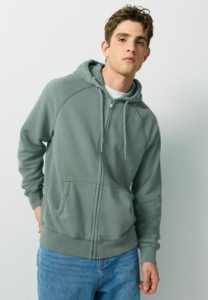 Young man wearing a muted green zip-up hoodie and blue jeans, standing with one hand behind his back against a plain light background.