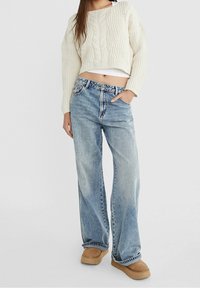 Suéter corto de punto trenzado blanco, pantalones vaqueros de pierna ancha en azul claro y botas de tobillo beige. El suéter tiene un patrón texturizado; los vaqueros presentan un lavado desgastado.
