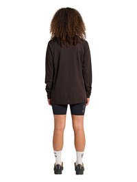 Personne aux cheveux bouclés debout, portant un t-shirt noir à manches longues, un short de cyclisme noir avec le texte "ATW", des chaussettes blanches et des baskets noires.