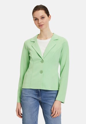 Blazer vert clair en tissu doux, avec un col cranté, deux boutons et une coupe cintrée, porté avec une chemise blanche en dessous.