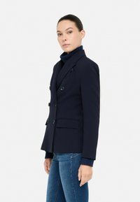 Námornícky dvojoblúkový blazer s dvoma veľkými prednými vreckami a čiernymi gombíkmi, kombinovaný s textúrovaným námorníckym rolákom. Hladký povrch látky.