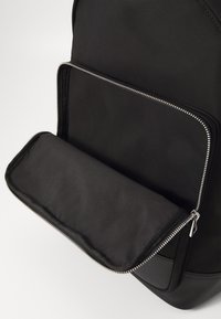 Zaino in tessuto nero con tasca frontale con zip. Presenta una superficie liscia e dettagli in metallo argentato. Design semplice e funzionale.