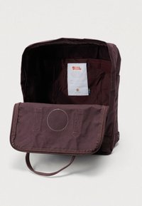 Bordowy plecak z tkaniny z przednią klapką, dużą otwartą przegrodą i metką wewnątrz. Posiada górny uchwyt oraz kontrastujące detale przeszyć.
