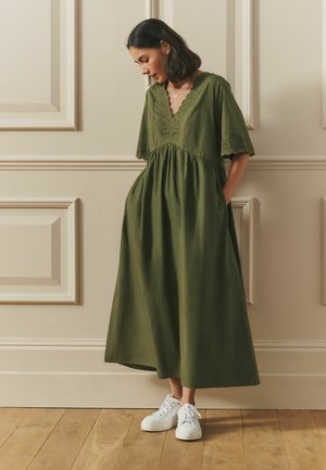 Olive green maxi dress with a scalloped V-neckline and lace detailing. Loose fit, short sleeves, and side pockets. Paired with white sneakers.