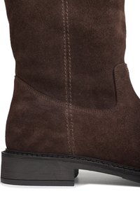 Botte en suede marron hauteur genou avec une surface texturée, des détails cousus et un petit talon noir. Présente une ouverture sur le côté.