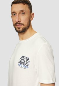 Witte katoenen t-shirt met een bedrukte graphic in zwarte en blauwe tekst aan de bovenste linkerzijde, ronde halslijn en korte mouwen.