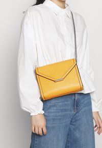 Osoba v bílé košili s knoflíky a modrých džínách, která nese malou žlutou kabelku přes rameno se stříbrným řetízkem.