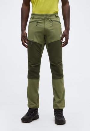 Mężczyzna w dwutonowych zielonych spodniach trekkingowych z zamykanymi kieszeniami i czarnych butach trekkingowych, stojący na jasnym tle.