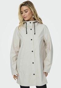 Veste imperméable beige clair avec capuche, cordons noirs et boutons-pression. Dotée de poches latérales et d'une coupe décontractée. Texture lisse.