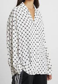 Camicia bianca con maniche lunghe caratterizzata da un motivo geometrico nero di catene e cuori, con una vestibilità rilassata e colletto a bottoni.
