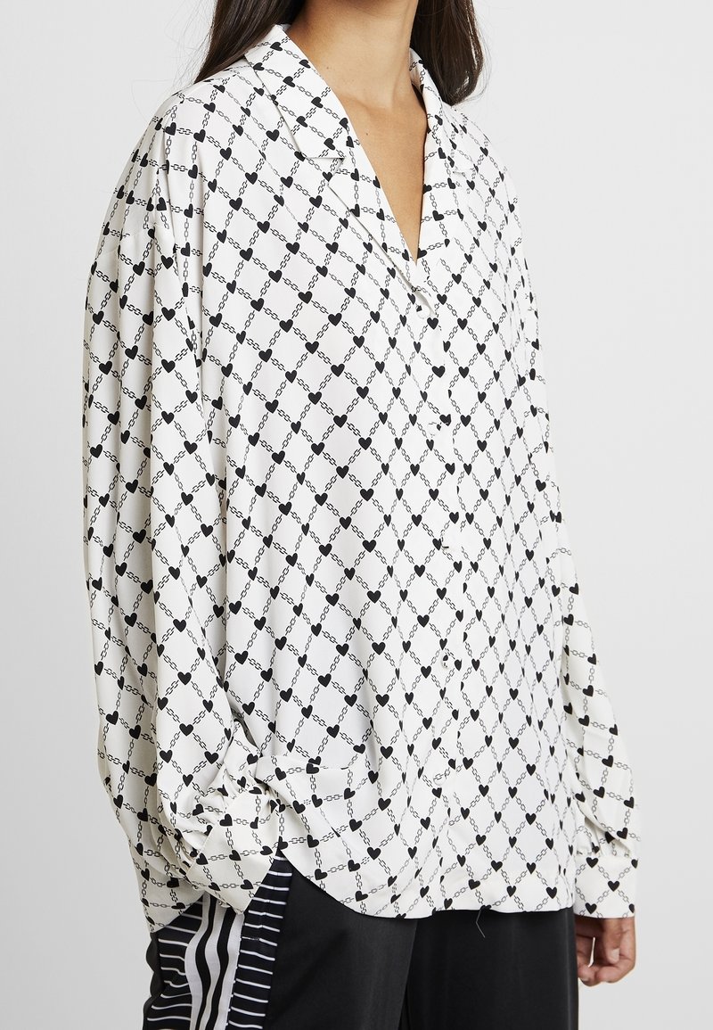 Camicia bianca con maniche lunghe caratterizzata da un motivo geometrico nero di catene e cuori, con una vestibilità rilassata e colletto a bottoni.