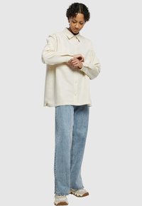 Camicia button-up beige chiaro con maniche lunghe, vestibilità rilassata e spacchi laterali, abbinata a jeans blu a gamba dritta e sneakers crema.