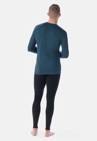 Long-sleeve teal shirt with a smooth texture, paired with fitted black leggings. Model shown from the back, barefoot, against a white background.