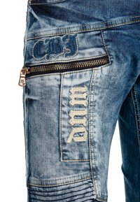 Denim-Cargo-Hosen in verblasstem Blau mit gesticktem Text "CBJ" und "WRC." Merkmale sind eine vordere Reißverschlusstasche und Rippendetails an den Beinen.