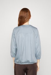 Blusa azul con estampado, de mangas tres cuartos, cuello redondo y corte holgado. Presenta un detalle de espalda fruncida y una textura suave.