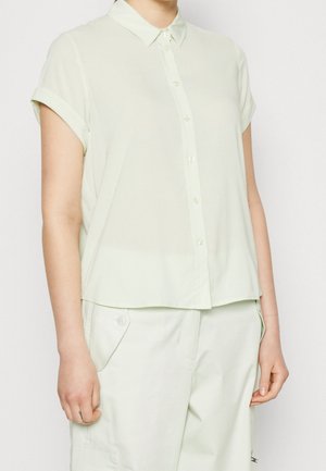 Frau trägt ein hellgrünes, kurzärmliges Hemd mit Kragen und passende Hose mit geknöpften Taschen.
