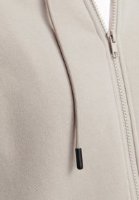 Sweat à capuche zippé beige avec une texture lisse, un embout noir sur le cordon de serrage et une doublure intérieure blanche contrastante.