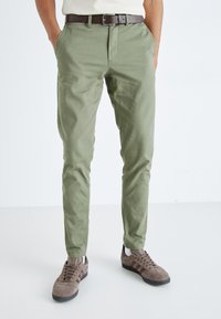 Chinos verde caqui hechos de tejido de algodón ligero, con cremallera, bolsillos laterales y un diseño ajustado, combinados con zapatillas marrones.