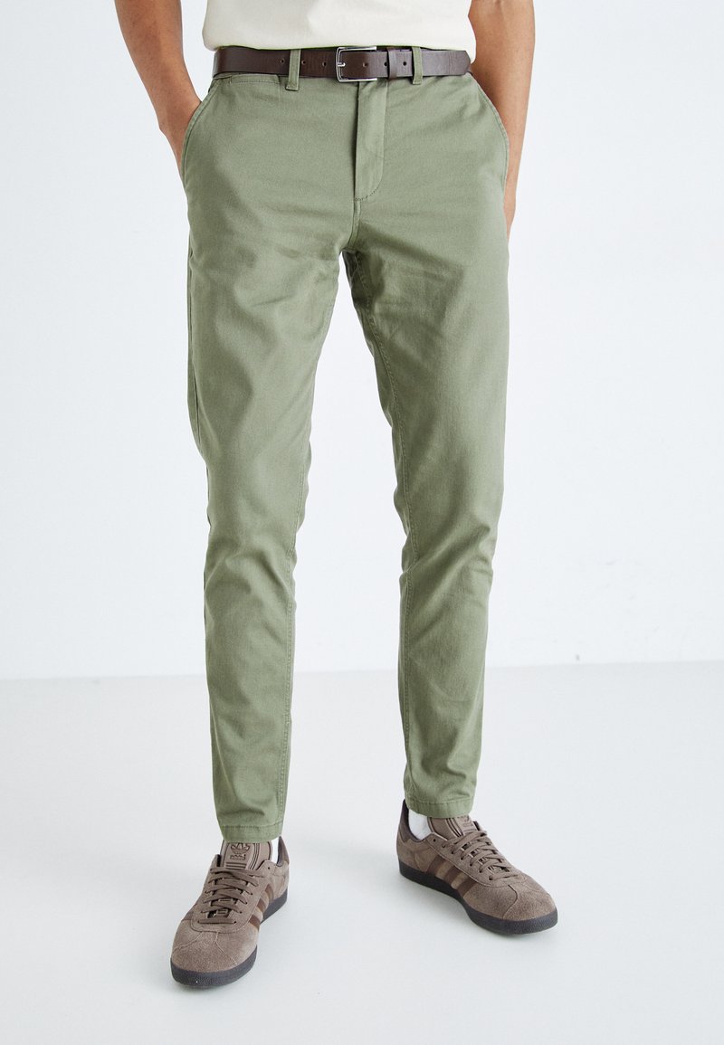 Chinos verde caqui hechos de tejido de algodón ligero, con cremallera, bolsillos laterales y un diseño ajustado, combinados con zapatillas marrones.