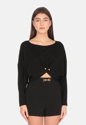 Top cropped nero con maniche lunghe, caratterizzato da un dettaglio attorcigliato nella parte frontale e accenti in metallo dorato. Abbinato a pantaloni corti a vita alta.