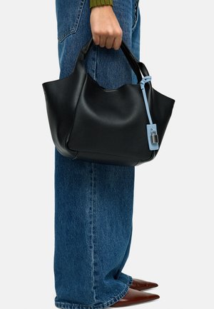 Osoba trzymająca dużą czarną skórzaną torbę na ramię z niebieską metką i srebrną kłódką, ubraną w niebieskie dżinsy i brązowe spiczaste buty.