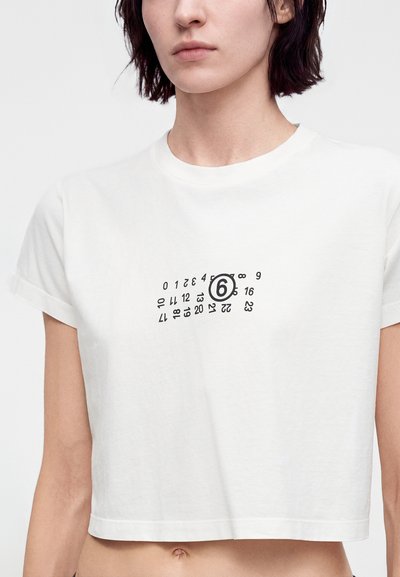 Γυναίκα που φοράει λευκό crop top με μαύρους αριθμούς ημερολογίου σκορπισμένους και τον αριθμό 6 κυκλωμένο στο κέντρο.