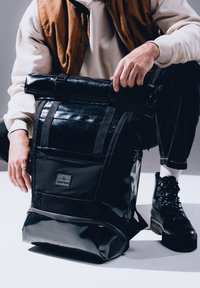 Fekete, feltekerhető hátizsák fényes anyagból, több pánttal, egy elülső zsebbel és márkás címkével. A textúrás elemek gazdagítják a dizájnt.